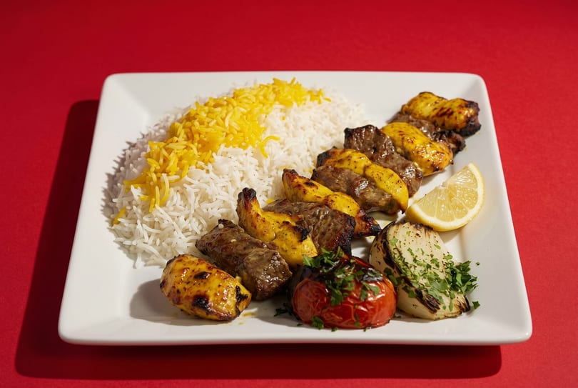 A white plate with rice, chicken and beef kabobs, grilled tomato, onion, and a lemon wedge.